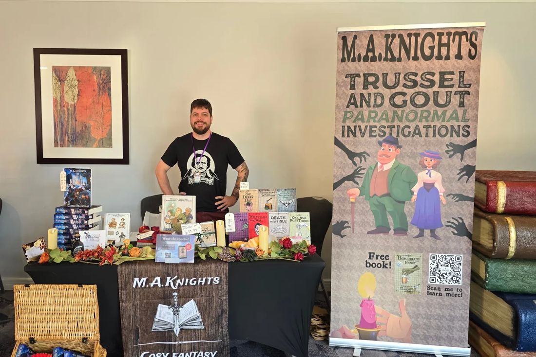M.A. Knights behind his signing table, covered in books, with Trussel and Gout banners